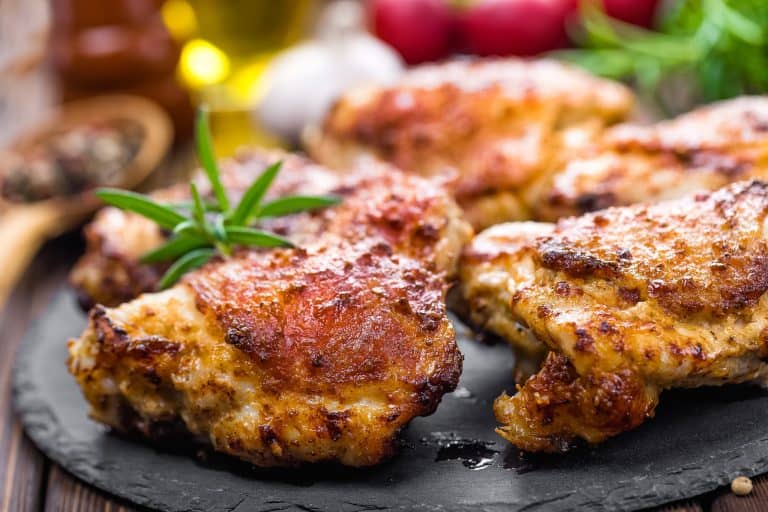Juicy grilled chicken thighs with crispy, golden-brown skin garnished with fresh herbs on a black slate serving platter. Perfect for barbecue dinners and savory meal options.