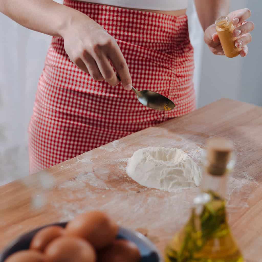 Scooping flour on wooden kitchen table with spoon, close-up; person adding peanut butter to baking ingredients, home kitchen; preparing dough for baking, baking ingredients on rustic wooden surface; image of baking preparation showing flour, peanut butter, eggs; relevant for home baking, cooking, kitchen essentials, baking supplies, food preparation.