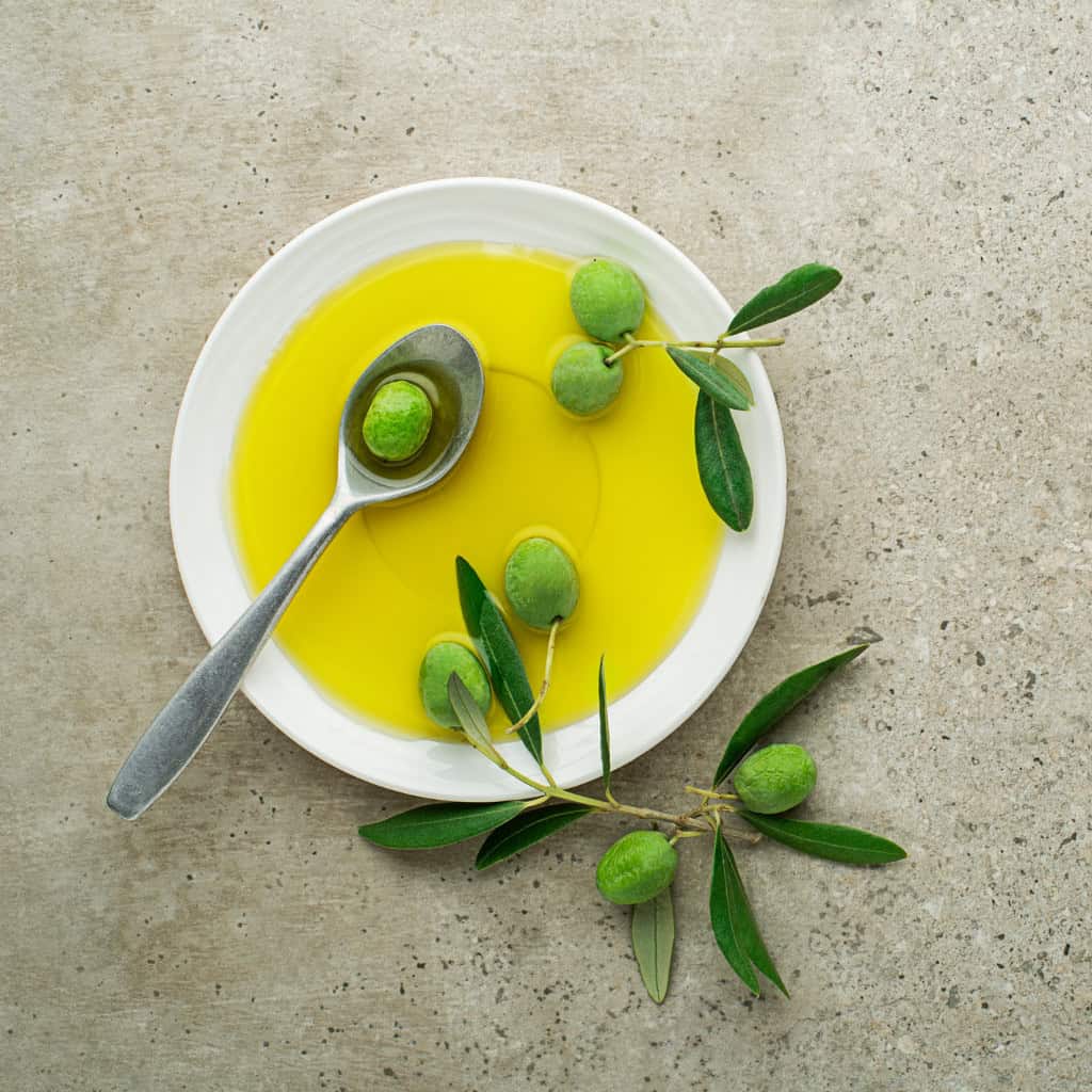 Fresh green olives in olive oil with a silver spoon and olive branch on a neutral background.