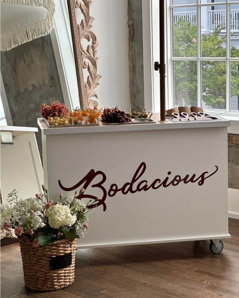 Vibrant fruit and cheese display on a white mobile serving cart with "Bodacious" logo in a stylish, rustic space with large windows and floral decor.