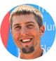 Portrait of a smiling person with short light brown hair and a short beard, set against a colorful background with indistinct text, capturing the warmth and satisfaction often found in thriving HVAC careers.
