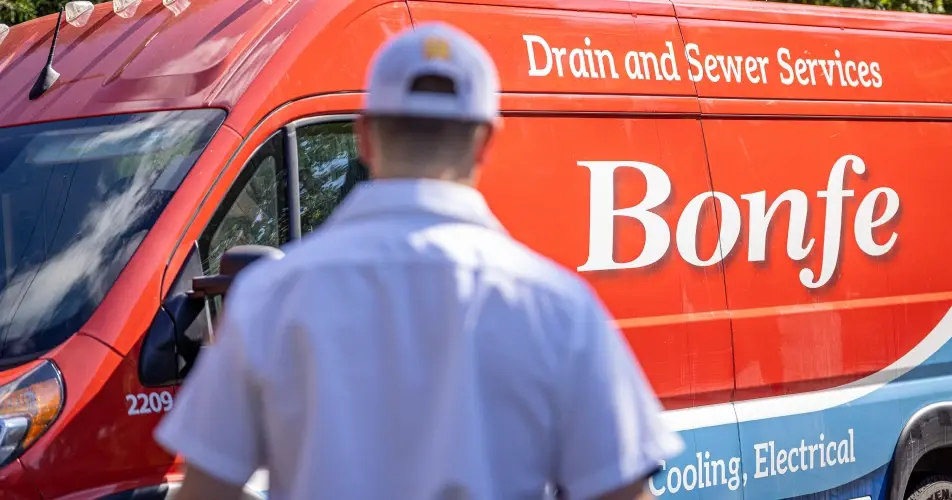 A person in a white cap and uniform stands in front of a red Bonfe service van with "Drain and Sewer Services" and "Cooling, Electrical" written on it.
