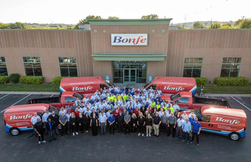 A large group of employees stands proudly in front of the building marked "Bonfe," embodying the spirit captured in our "About Us" ethos. Four branded vans are neatly parked out front, symbolizing our commitment and teamwork.