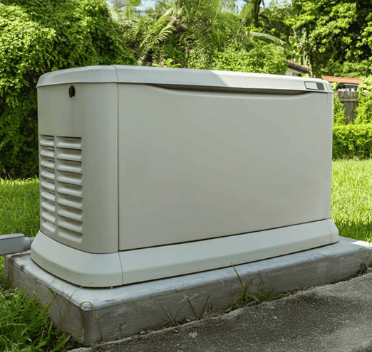 A gray generator installation is set on a concrete slab in a grassy yard, enveloped by lush greenery and trees.