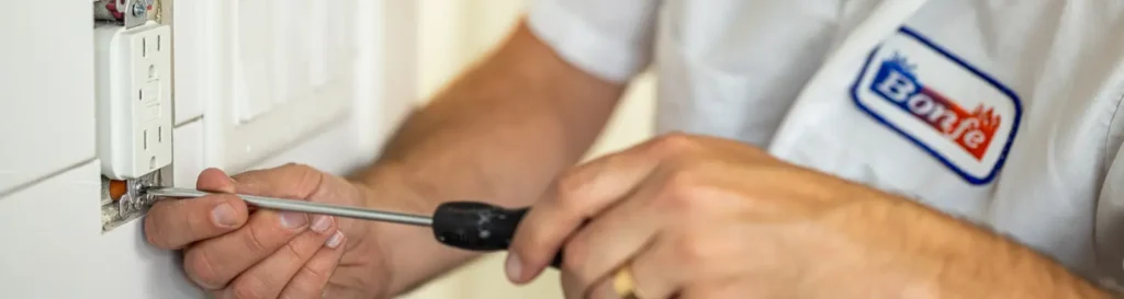 A person wearing a "Barle" uniform uses a screwdriver to install or repair a wall electrical outlet.