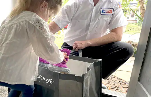 A service worker kneels at a doorway next to a young girl, placing boots into a shopping bag—a small act of kindness reflecting the spirit of community in Minnesota.