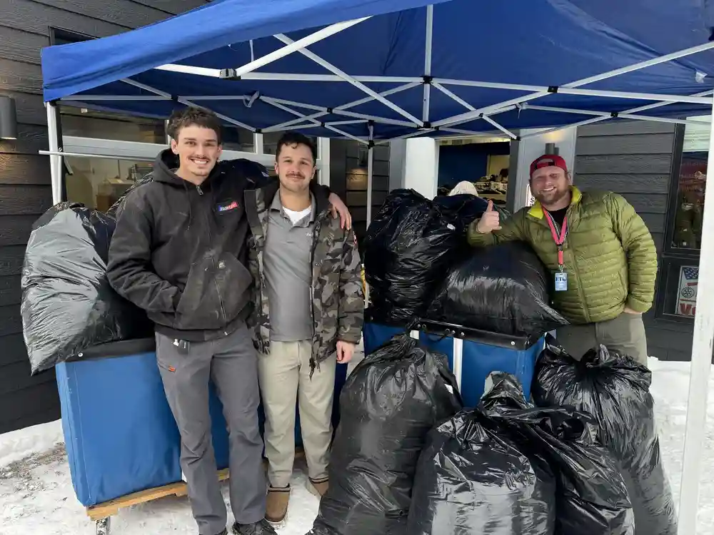 Three men stand under a blue canopy outdoors, showcasing Bonfe's community involvement as they pose with large black garbage bags placed in blue bins on a snowy surface.