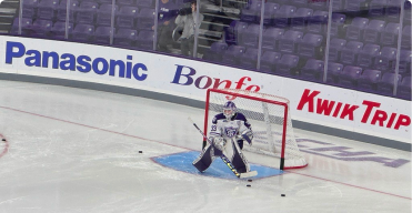 A hockey goalie in full gear stands in front of the goal net on an ice rink during practice, with three pucks nearby and Bonfe advertisements on the boards, highlighting community involvement in local sports.