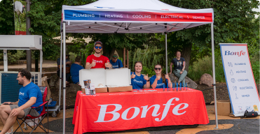 Three people sit and stand at a Bonfe-branded booth under a canopy, giving thumbs up. The booth displays promotional materials and a sign listing services, highlighting Bonfe's community involvement at local events.