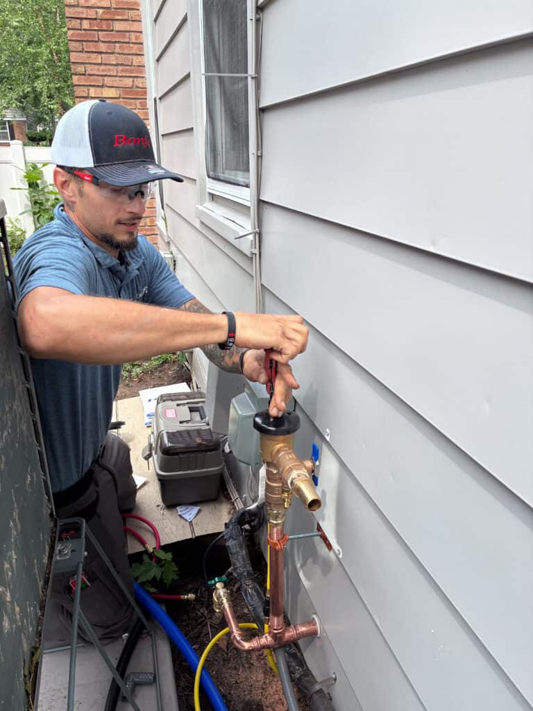 A person wearing safety glasses installs or repairs outdoor plumbing next to a house, using a pipe wrench on a brass fixture. Tools and equipment are nearby, capturing the hands-on expertise featured in the Bonfe Insider, August 2026 edition.