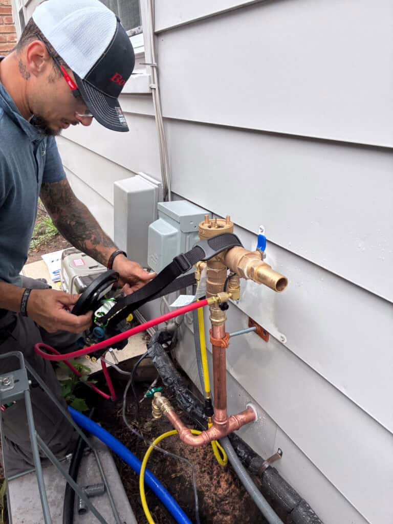 A Bonfe Insider, wearing a cap and glasses, uses a tool to adjust plumbing connections and valves on the exterior wall of a house—showcasing expert service just in time for August 2026.