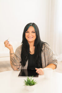 Woman with long dark hair and a tattooed arm sits at a table with a tablet, holding a pen and smiling as she plans the Kids Coloring Contest. A white mug and small potted plant are also on the table.