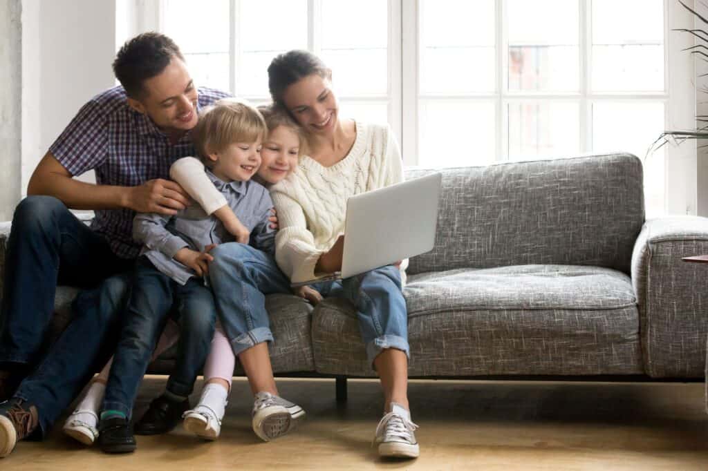 A family of four sits on a gray couch at home, smiling and looking at a laptop together in front of a large window, researching how to prepare their home for winter as colder weather approaches.