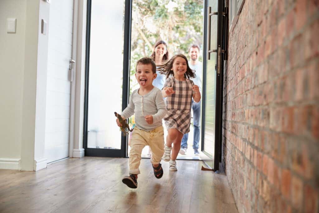 Two young children run excitedly through an open front door, with two adults smiling in the background—happy moments made possible when you keep your home warm and save money on heating.