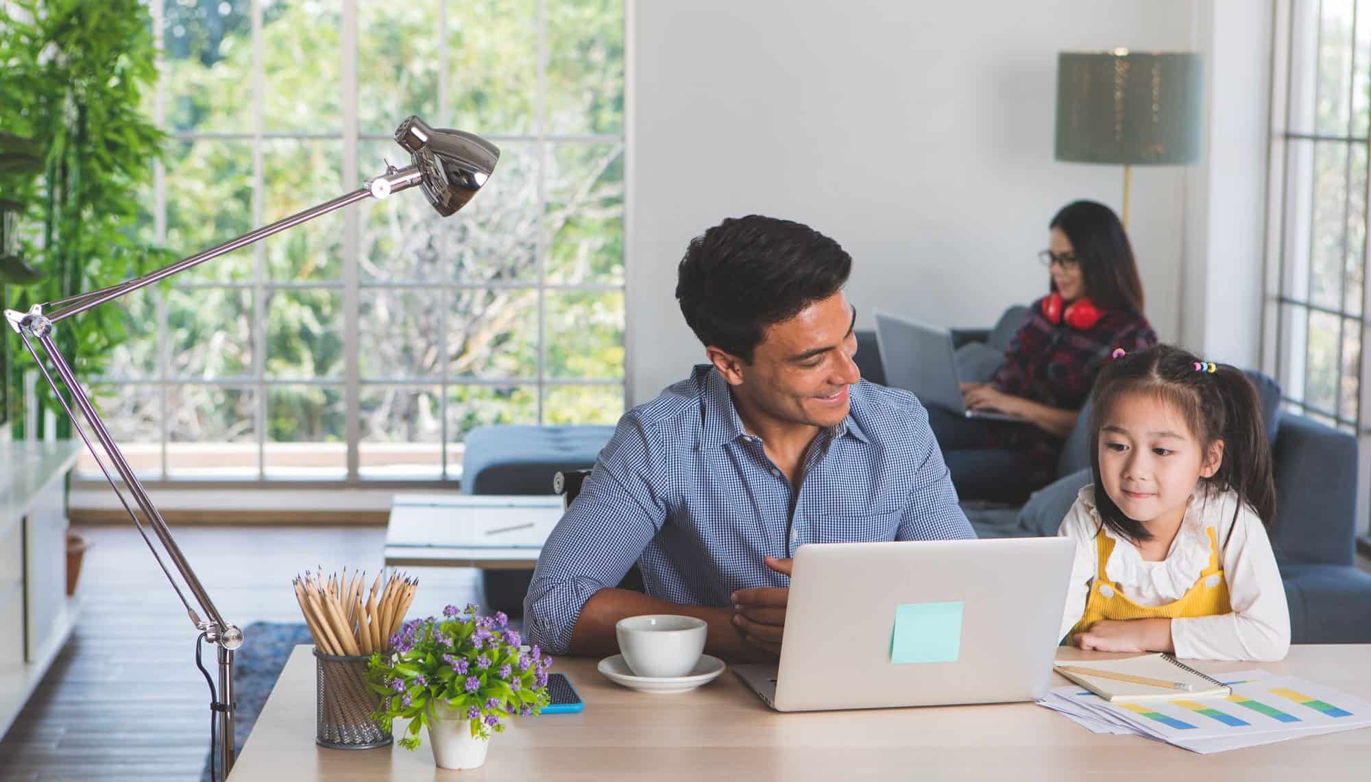 A man works on a laptop at a desk with a young girl beside him, while a woman uses her laptop on the couch in the background, showcasing home efficiency and comfort while working at home.