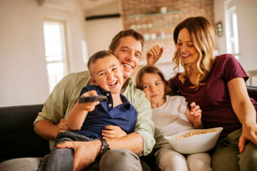 A family of four sits on a couch, smiling and eating popcorn while watching TV, enjoying the comfort and energy savings of their new high-efficiency HVAC system. The boy in front holds a remote and points it toward the camera.