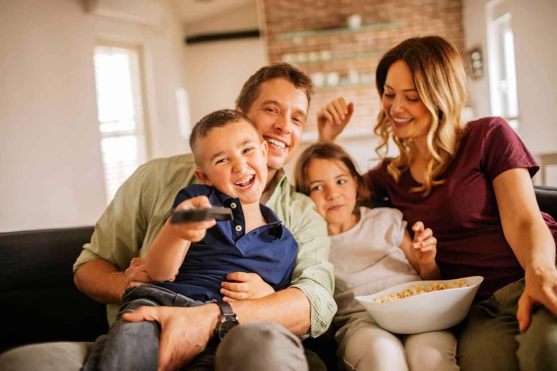 A family of four sits on a couch, smiling and eating popcorn while watching TV, enjoying the comfort and energy savings of their new high-efficiency HVAC system. The boy in front holds a remote and points it toward the camera.