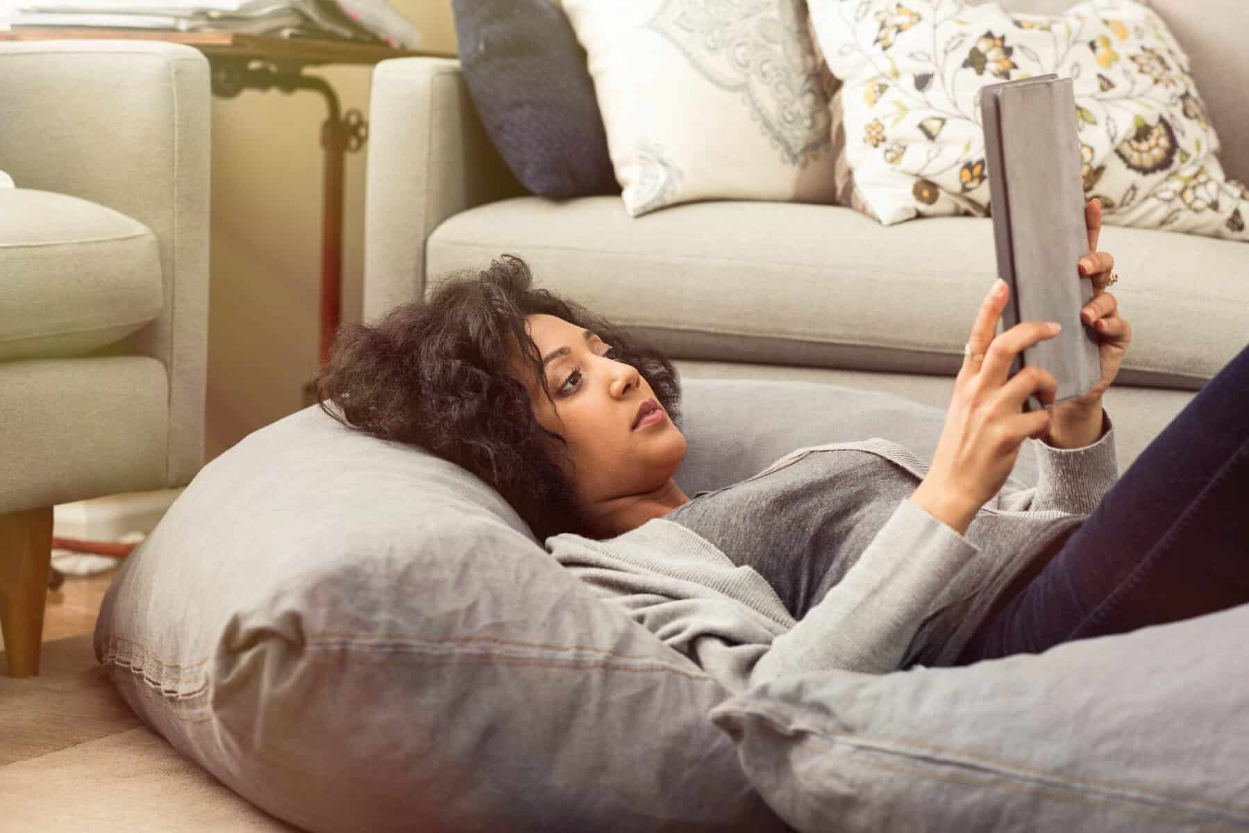 A woman lies on a large cushion in a living room, holding and looking at a tablet—she’s researching AC upgrade considerations for her home's air conditioning system.