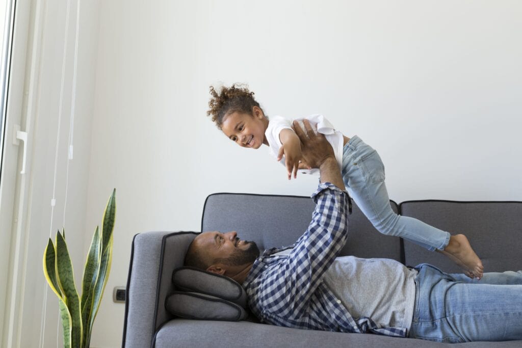 An adult lies on a sofa, lifting a smiling child above their chest near a potted plant, enjoying family time in the comfort of an efficient home heating and cooling system.