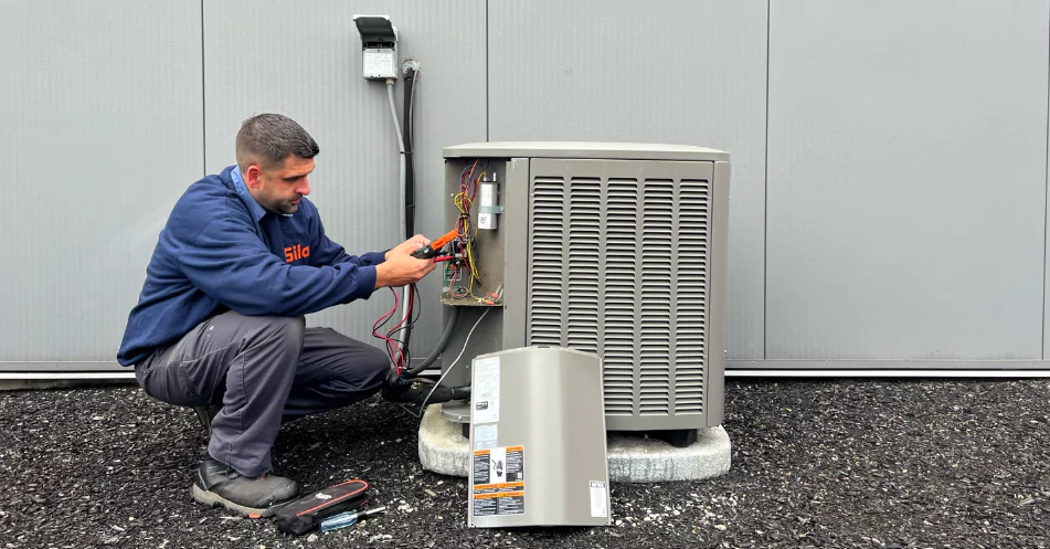 A technician kneels beside an outdoor HVAC unit, using tools to work on its wiring with the panel cover removed and placed on the ground.