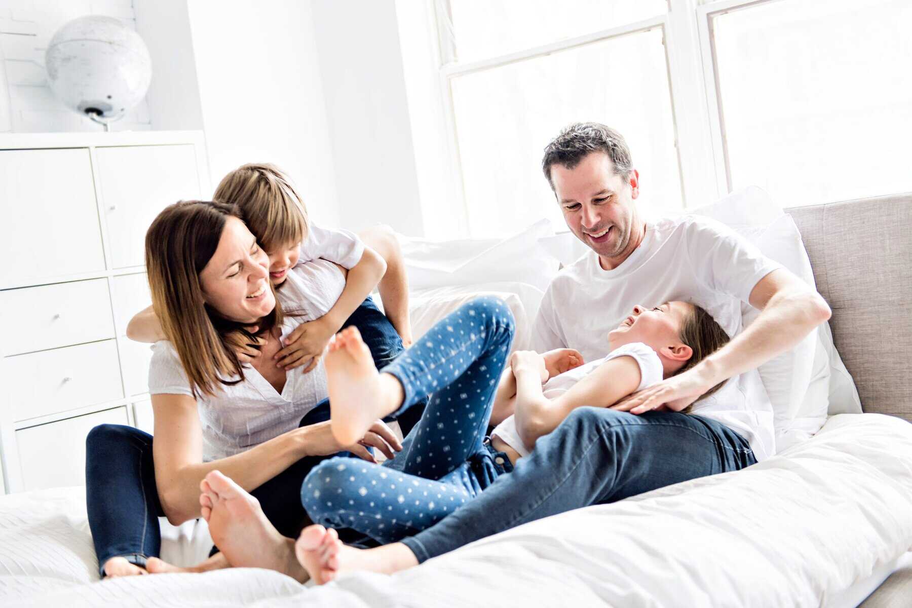 A family of four, two adults and two children, sit on a bed in a bright room, laughing and playing together while enjoying cool air from the AC.