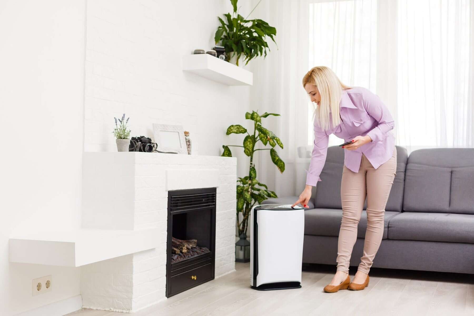 A woman in a light purple blouse presses a button on an air purifier with humidity control in a modern living room featuring plants, a fireplace, and a gray sofa.