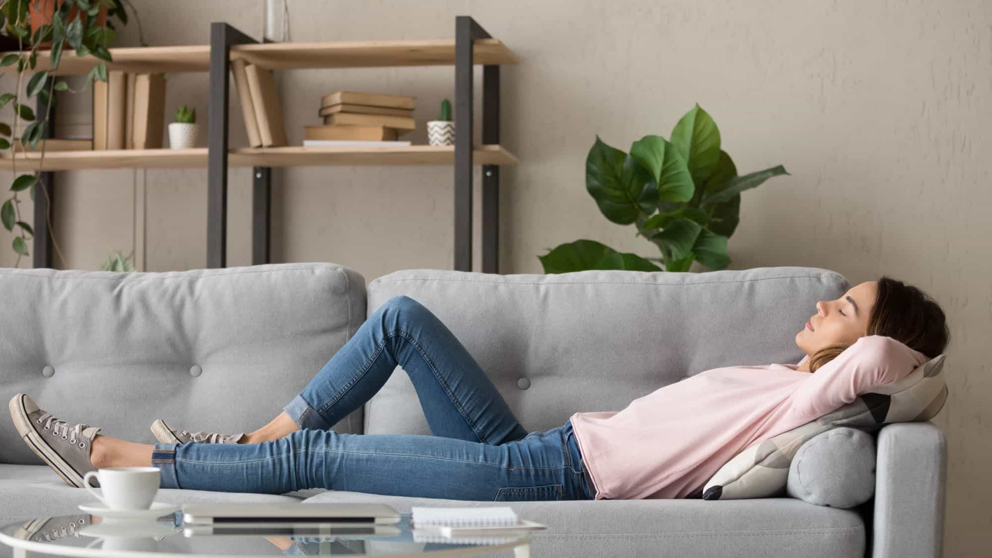 A woman in jeans and a pink top is lying on a gray sofa, relaxing in a living room filled with potted plants—one of the most effective ways to improve air quality and boost home air quality naturally.