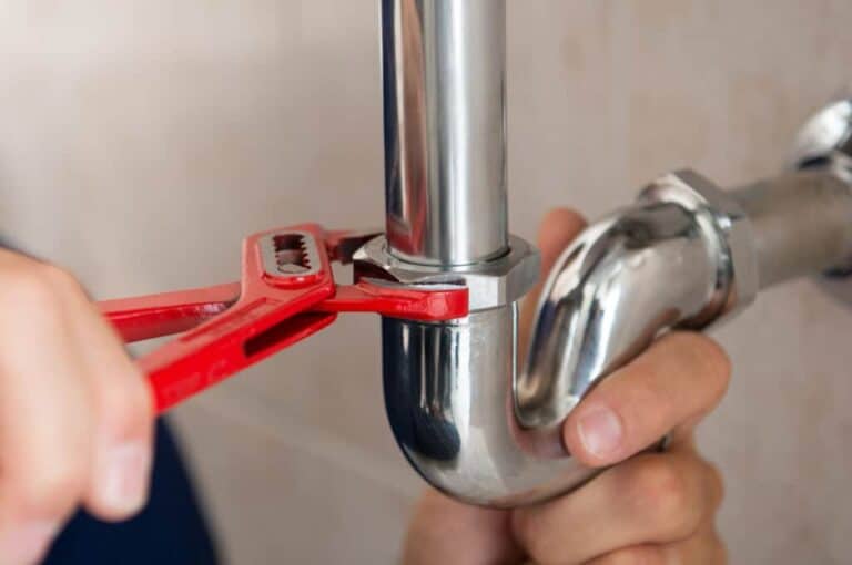 A person uses a red pipe wrench to tighten a metal pipe connection under a sink.