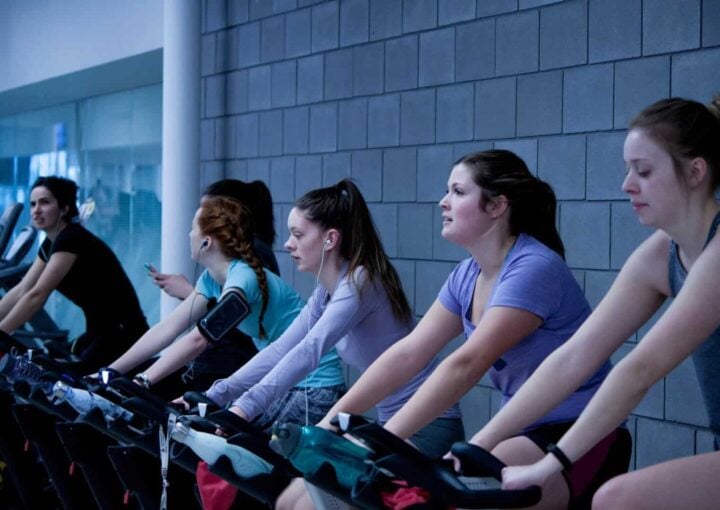 Indoor cycling class at Bay-O-Vista Swim & Tennis Club, featuring women exercising on stationary bikes with attentive instructors, promoting fitness and community engagement.