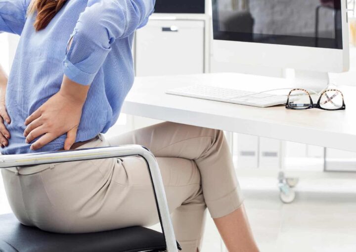 Woman experiencing lower back pain while sitting at her desk, highlighting health risks associated with prolonged sitting during desk jobs.