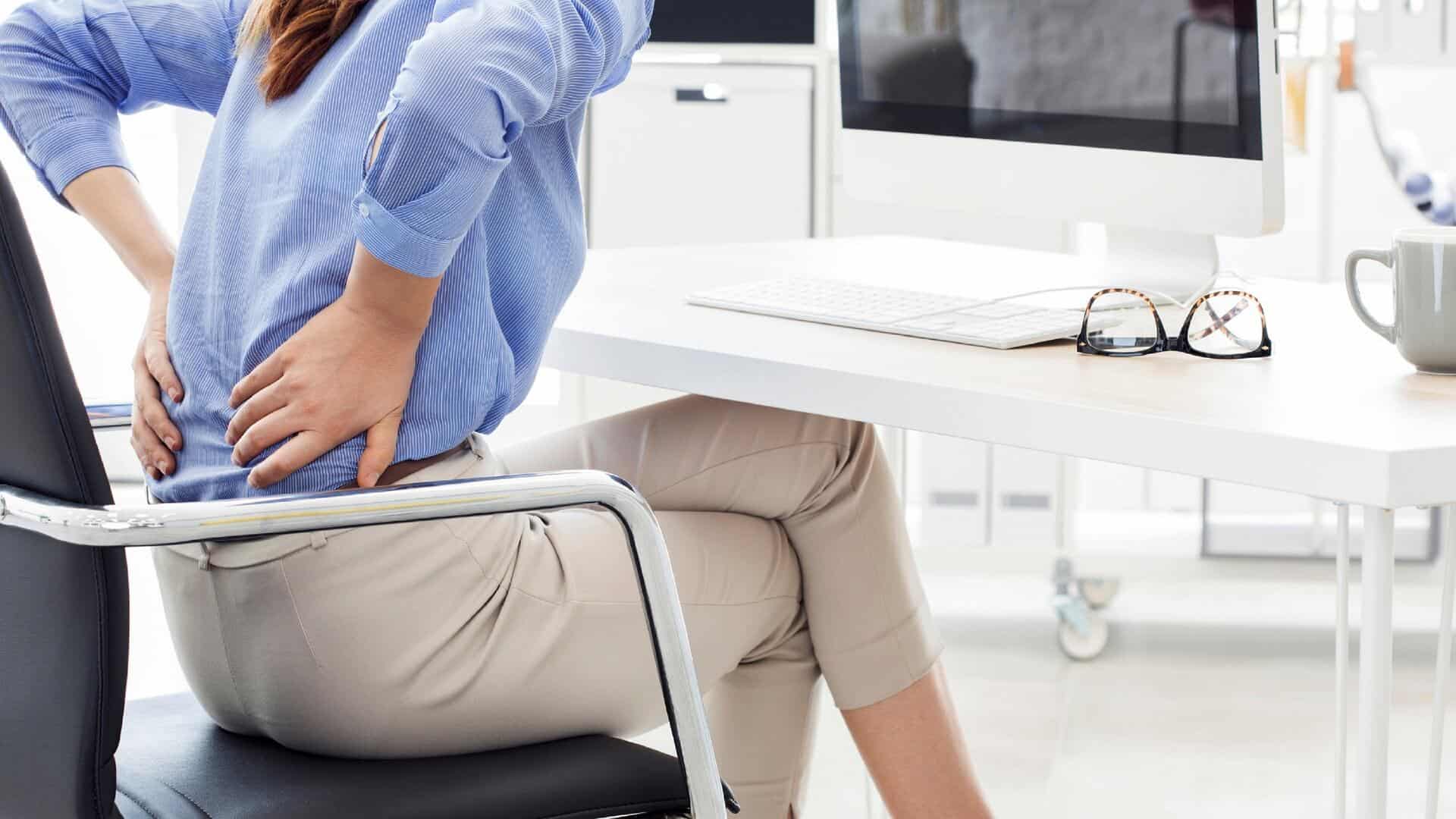 Woman experiencing lower back pain while sitting at her desk, highlighting health risks associated with prolonged sitting during desk jobs.