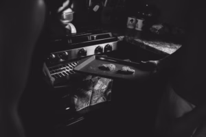 Two people are placing a baking tray with three pieces of food into an oven in a dimly lit kitchen, captured candidly by an Eau Claire wedding photographer.