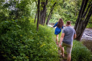 Two people are walking along a narrow, grassy path beside a river in a lush, green forest, captured beautifully by an Eau Claire wedding photographer.