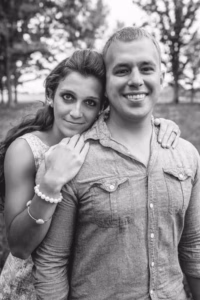 A woman stands behind a man, resting her hands on his shoulders. Both are smiling and posing outdoors with trees in the background, captured beautifully by an Eau Claire wedding photographer.