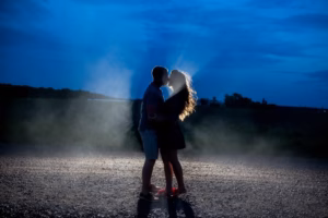 A couple stands close together on a gravel road at dusk, silhouetted against a blue sky with light shining behind them—captured beautifully by an Eau Claire Wedding Photographer.