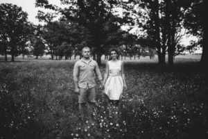 A man and woman stand holding hands in a field of wildflowers with trees in the background; captured in timeless black and white by an Eau Claire wedding photographer.