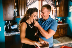 A man feeds a woman in a kitchen while both smile; the woman, mixing something in a bowl on the counter, shares this joyful moment—perfectly captured by an Eau Claire Wedding Photographer.
