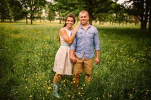 A woman and man stand together in a grassy field of yellow wildflowers, holding hands and smiling at the camera. Trees are visible in the background, captured beautifully by an Eau Claire Wedding Photographer.