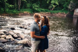 A man and a woman stand close together by a rocky riverbank, surrounded by trees and greenery, smiling at each other—captured beautifully by an Eau Claire Wedding Photographer.