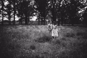 Two people walk together through a grassy field with wildflowers, surrounded by tall trees, in a black and white photograph by an Eau Claire wedding photographer.