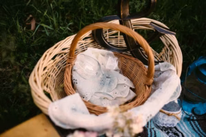 A wicker basket filled with lace-trimmed cloths, clear glassware, and a decorative black ampersand sits on grass—perfect inspiration from an Eau Claire Wedding Photographer.