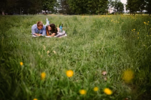 A man and woman lie on a blanket in a grassy field with wildflowers, reading and talking, with picnic items nearby—perfect inspiration for an Eau Claire Wedding Photographer capturing candid romance.