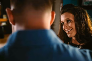 A woman with brown hair smiles while facing a man whose back is to the camera. Captured by an Eau Claire wedding photographer, the intimate moment unfolds in a cozy indoor setting.