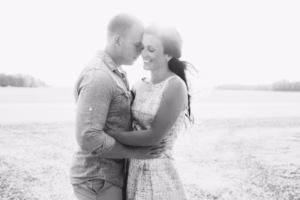 A couple stands close together outdoors, smiling and embracing, with sunlight shining behind them in a black and white photo captured by an Eau Claire Wedding Photographer.