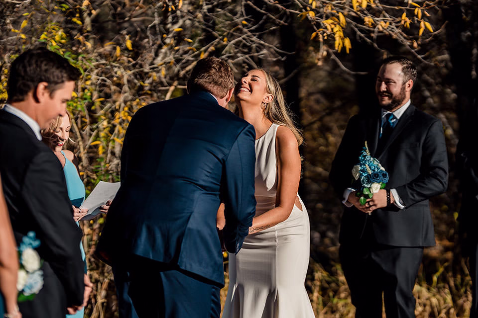 A bride and groom smile and lean in for a kiss during their Silverthorne Pavilion Wedding ceremony, with bridesmaids and groomsmen standing nearby.