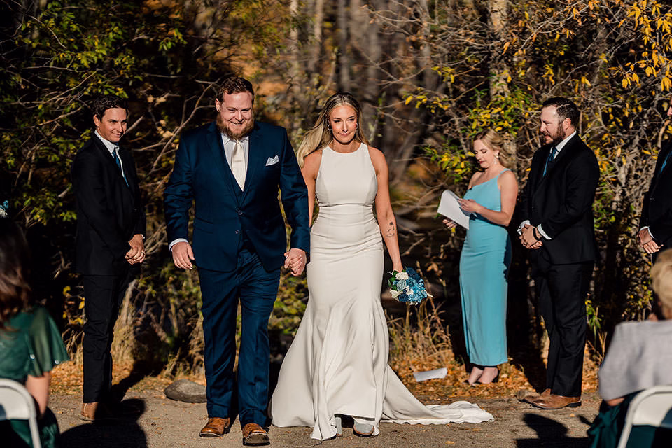 A bride and groom walk hand in hand outdoors after their Silverthorne Pavilion Wedding, with attendants and guests standing nearby among trees.