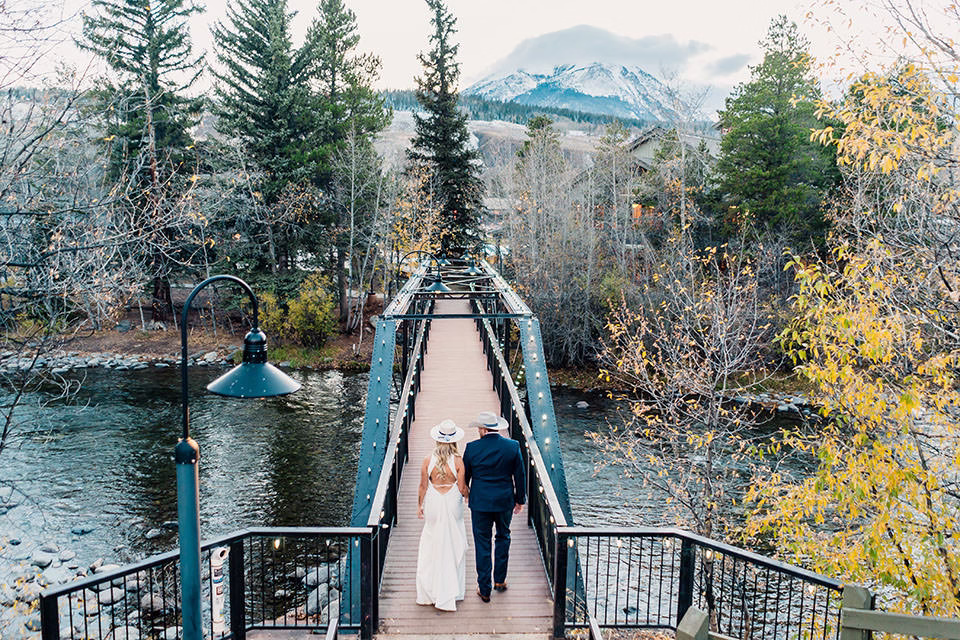 A couple in formal attire walks hand in hand across a wooden bridge over a river at their Silverthorne Pavilion Wedding, surrounded by autumn trees and mountains in the background.