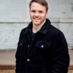 A man in a black jacket smiling, representing our company's enthusiasm and professionalism, posing in front of a brick wall.