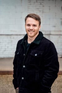 A man in a black jacket smiling, representing our company's enthusiasm and professionalism, posing in front of a brick wall.