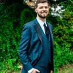 A groom in a tuxedo smiling in front of bushes.